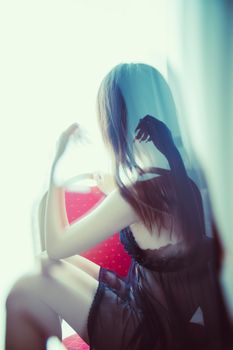 Artistic photograph of a woman in sheer lingerie seated subtly behind a curtain, creating a dreamy effect.