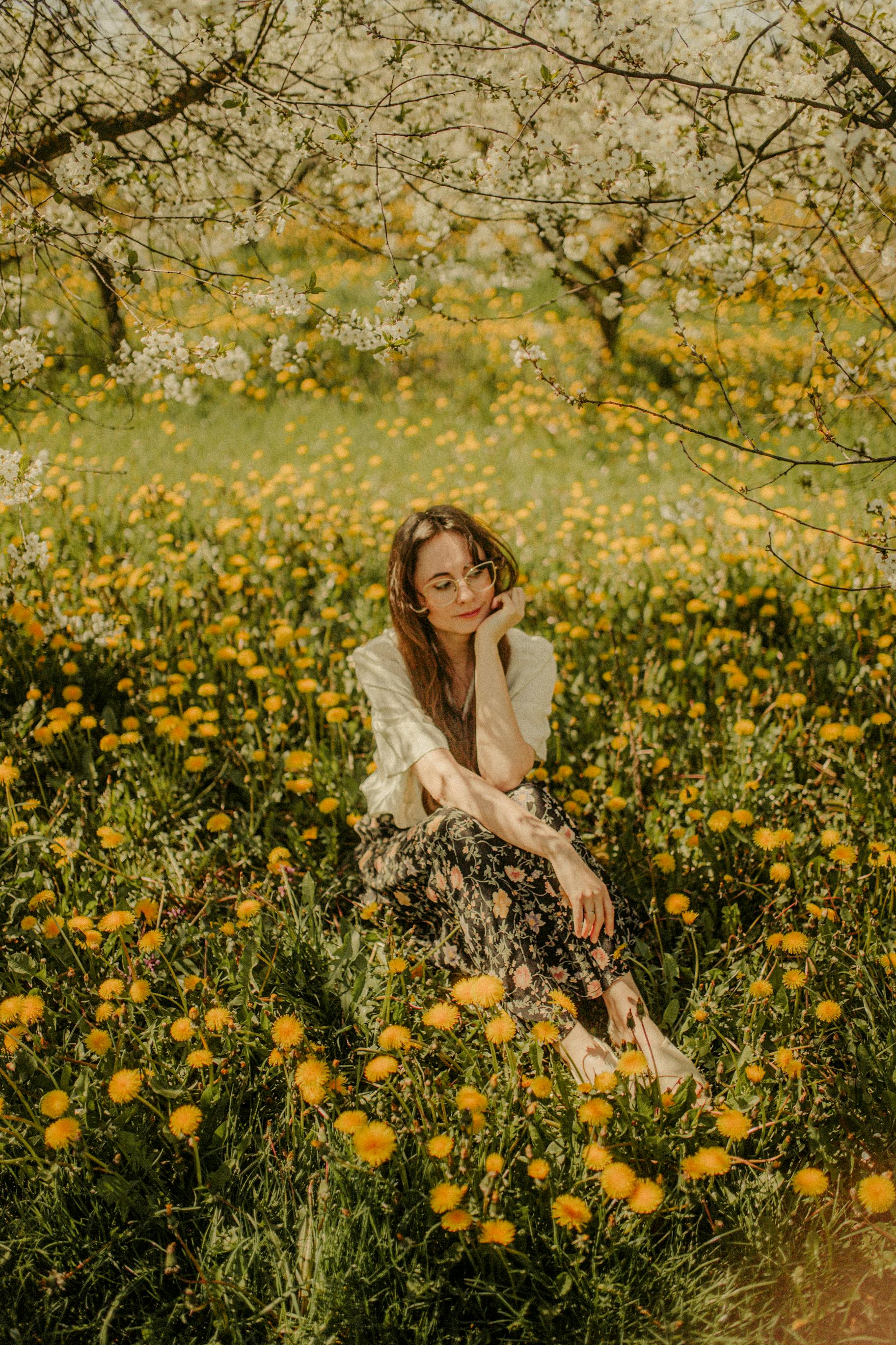 A young woman in a floral dress sits in a vibrant yellow wildflower meadow under blooming trees.