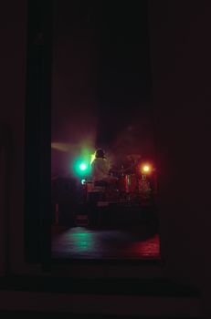 A drummer plays passionately on stage with colorful lights creating a dramatic atmosphere.