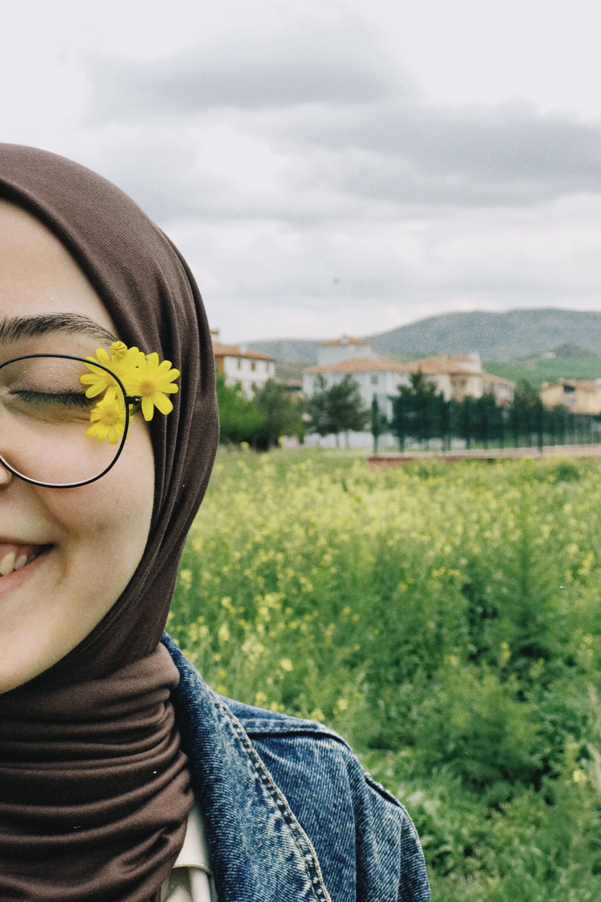 A joyful woman wearing a hijab and eyeglasses with a yellow flower, standing in a lush meadow.