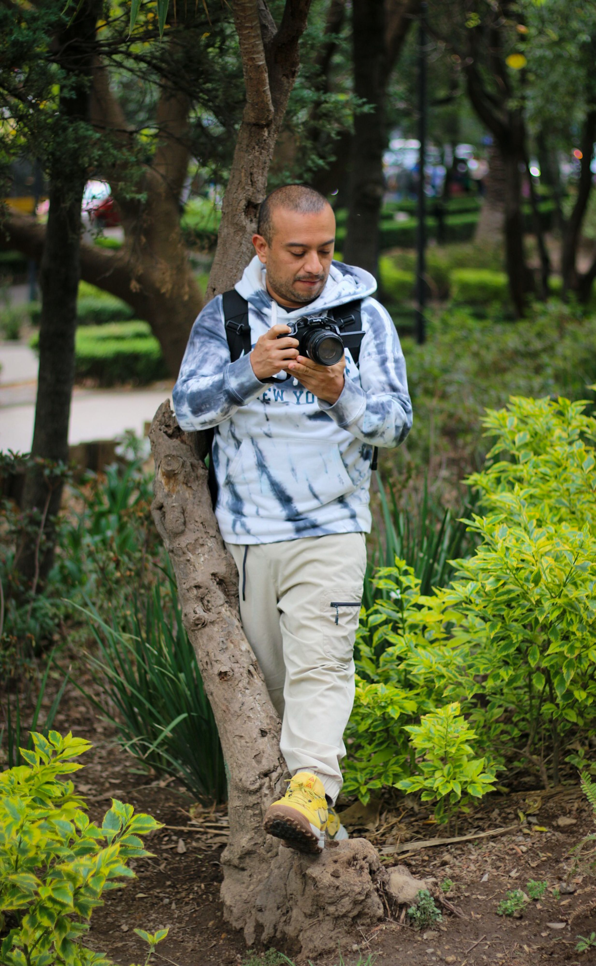 A man in a hoodie stands with a camera in a lush Ciudad de México park, capturing urban nature.