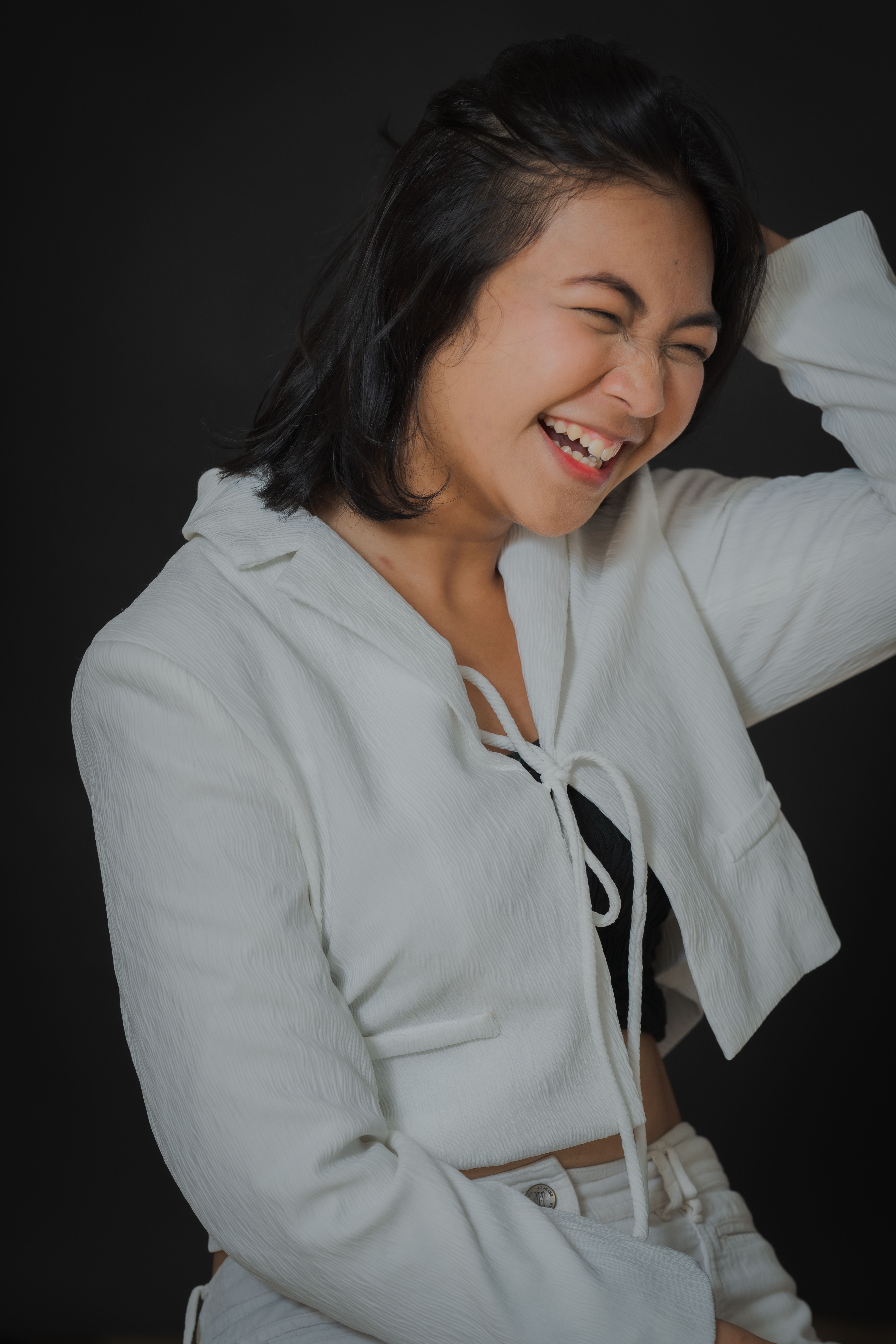 Young woman in white jacket laughing with hand in hair against a black background.