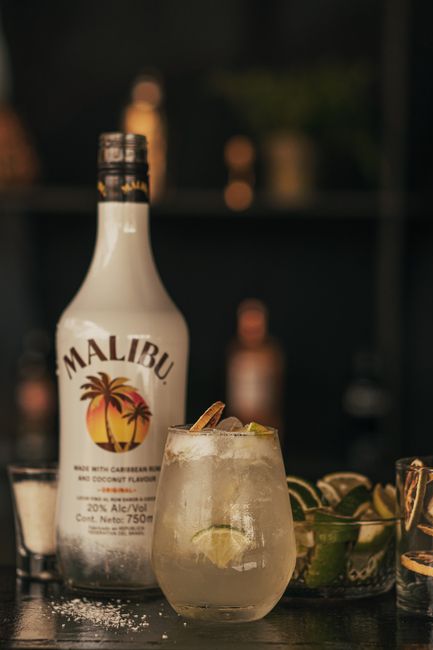 A tall glass of pale pink Malibu Bay Breeze cocktail with ice, garnished with a pineapple wedge and a maraschino cherry, on a bar towel next to a bottle of coconut rum.