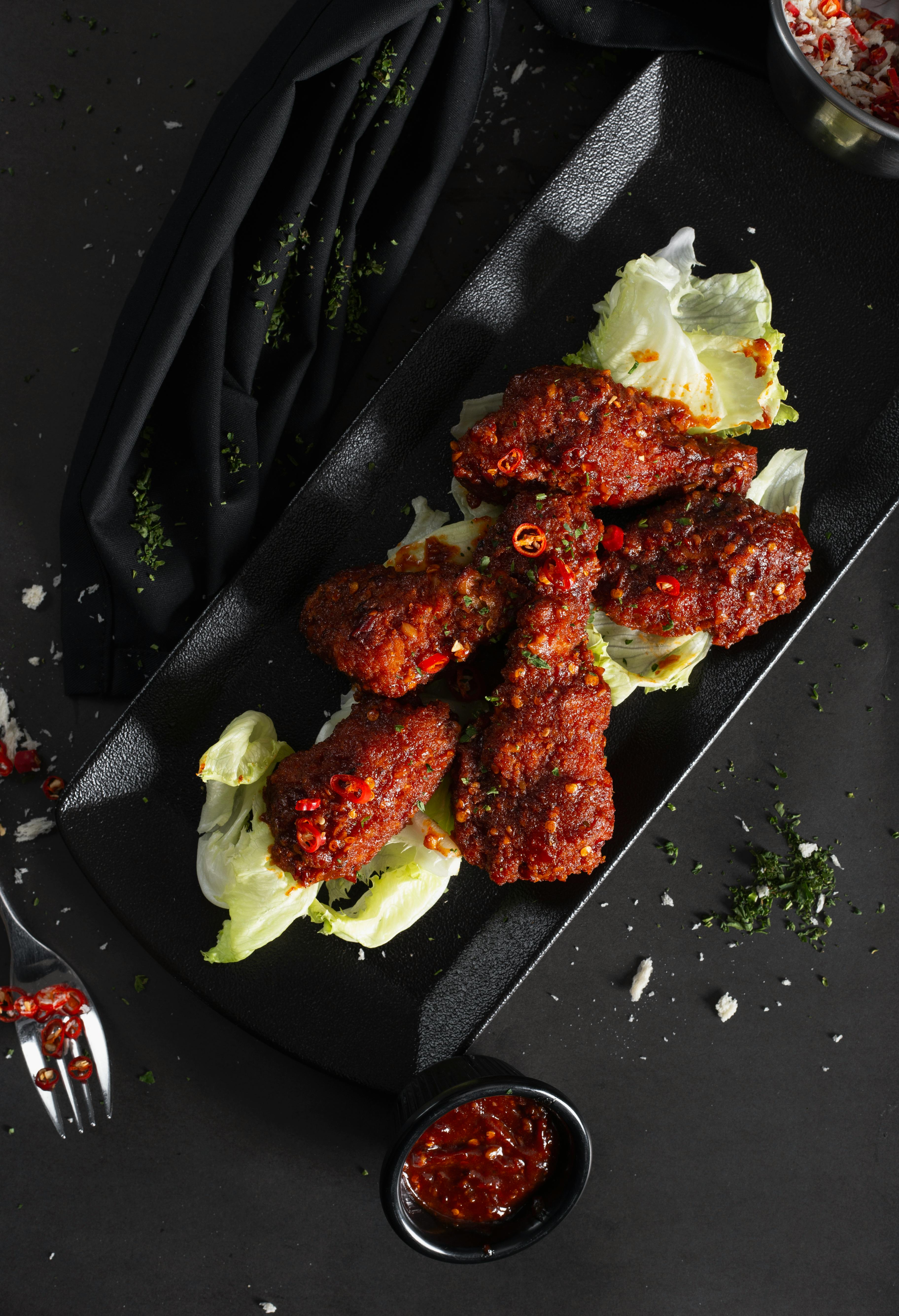 Delicious spicy chicken drumsticks on a black tray with fresh lettuce and dipping sauce.