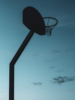 Silhouetted basketball hoop with net at twilight, creating a moody, dramatic scene.