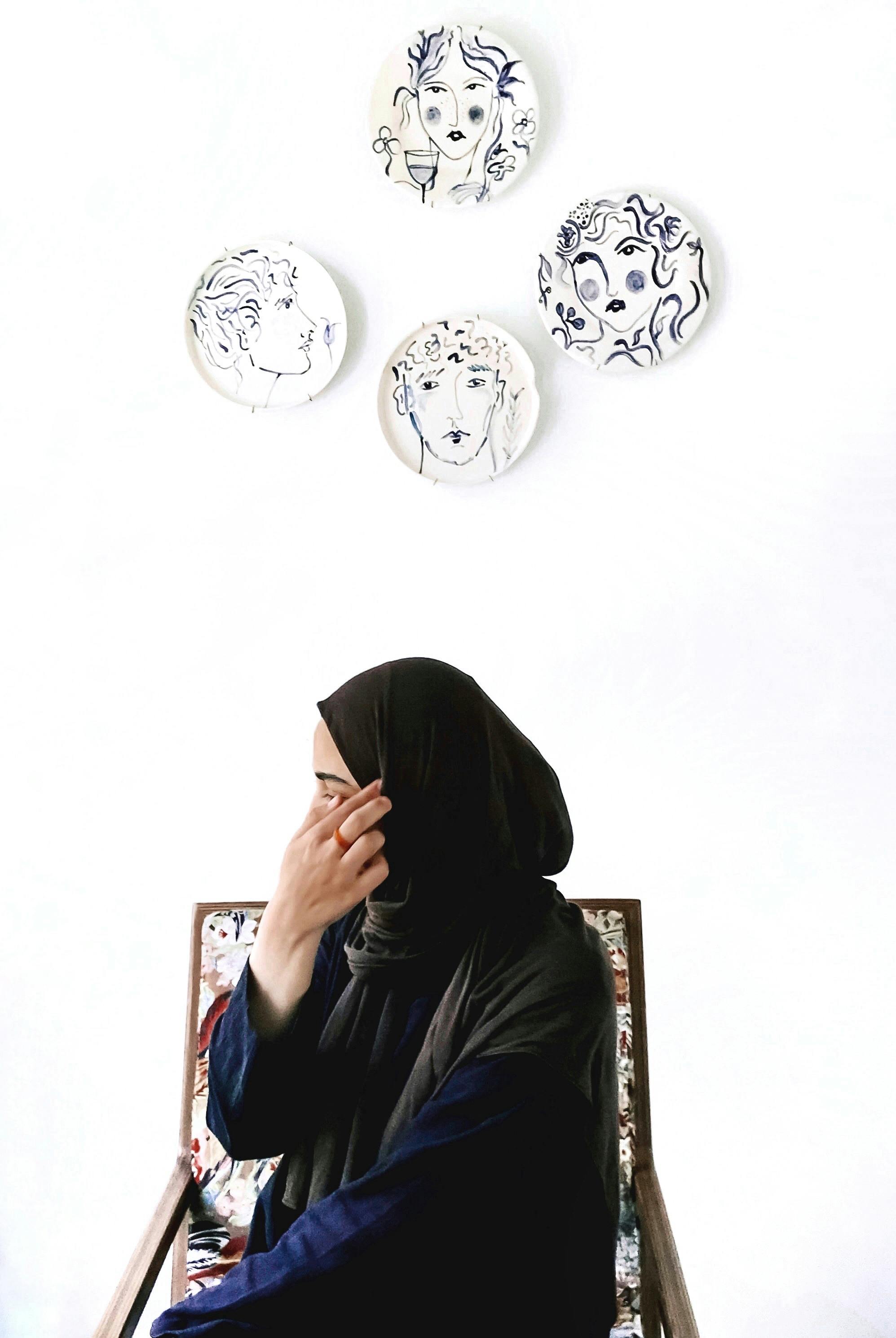 Woman in a hijab sitting in a chair against a white background with artistic wall plates.