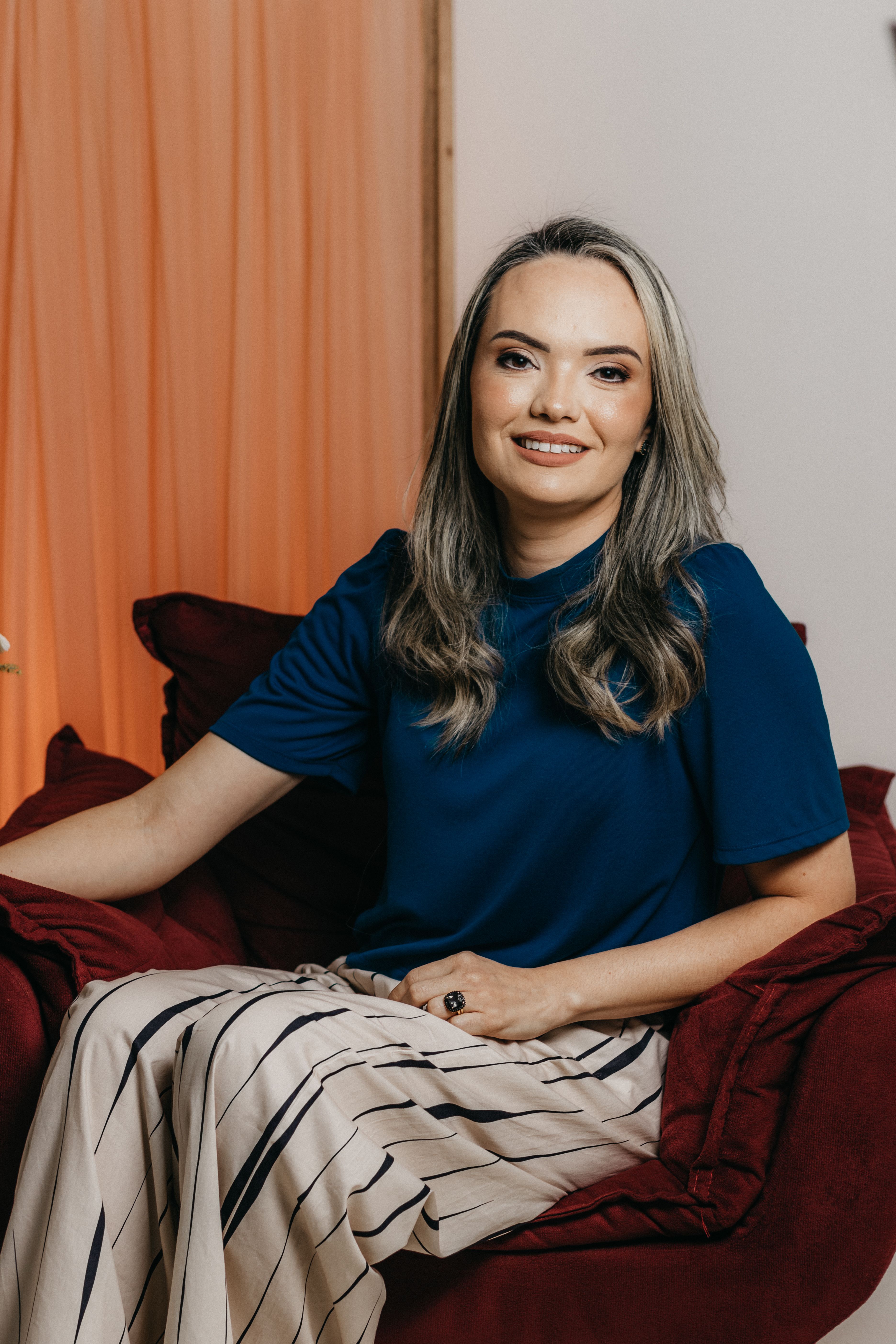 Elegant woman with gray hair and blue shirt sitting in a red armchair, indoors, smiling warmly.