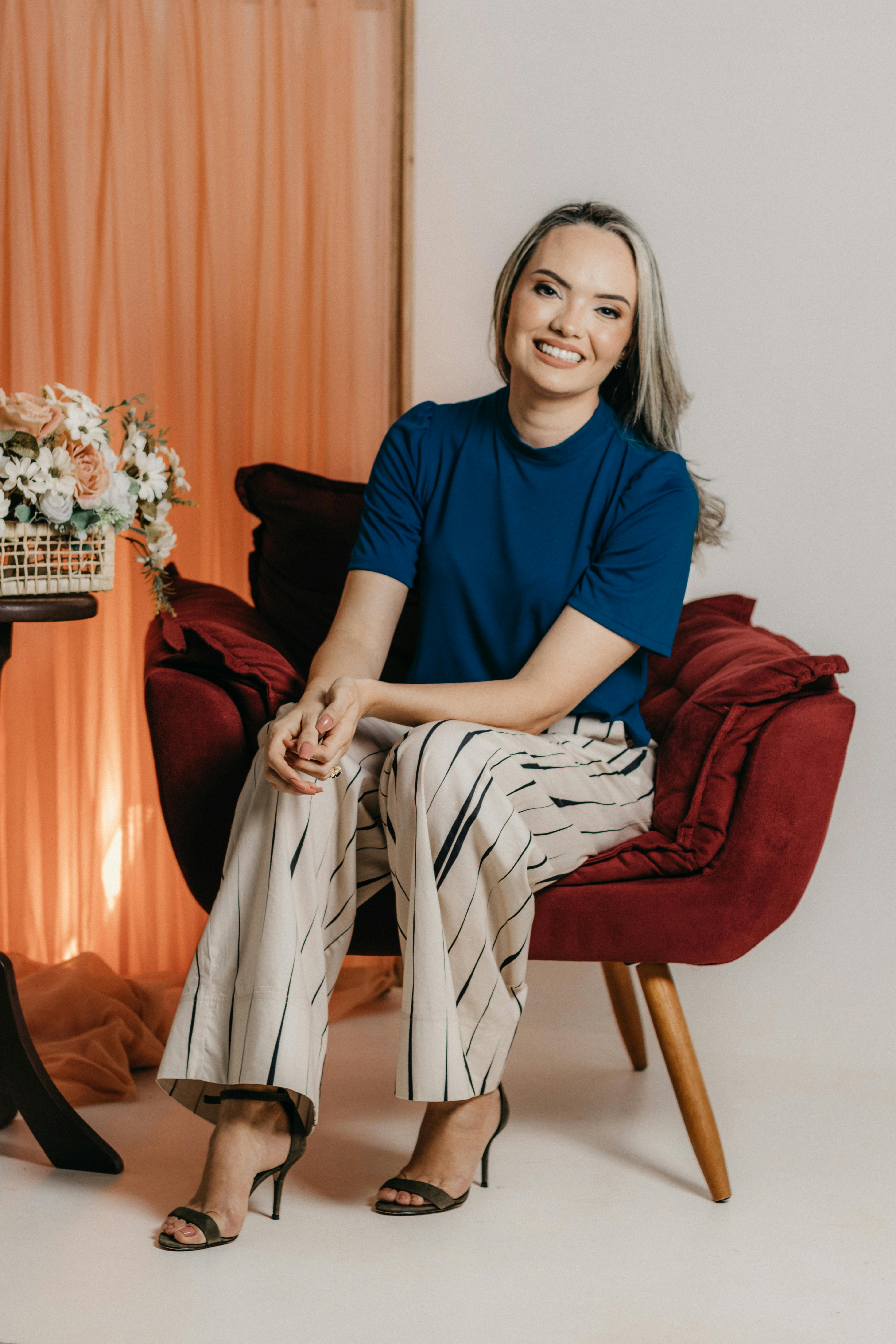 Blonde woman in a blue shirt and striped pants smiling while seated on a red armchair indoors.