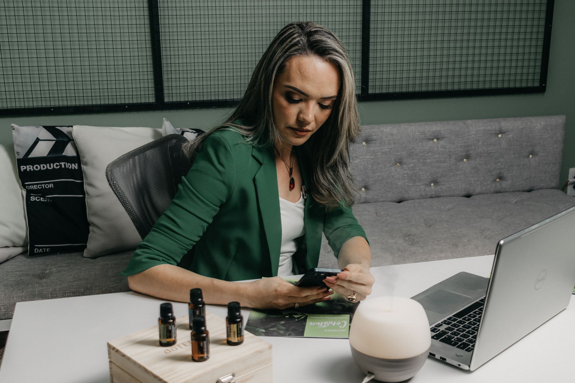 A businesswoman in a green jacket multitasks with a phone and laptop at a modern office desk.