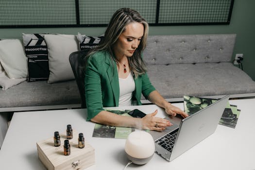 Woman in green suit working on laptop at home office, surrounded by essential oils and diffuser.