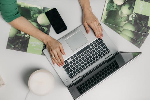 A woman with red nails types on a laptop, smartphone and documents nearby.