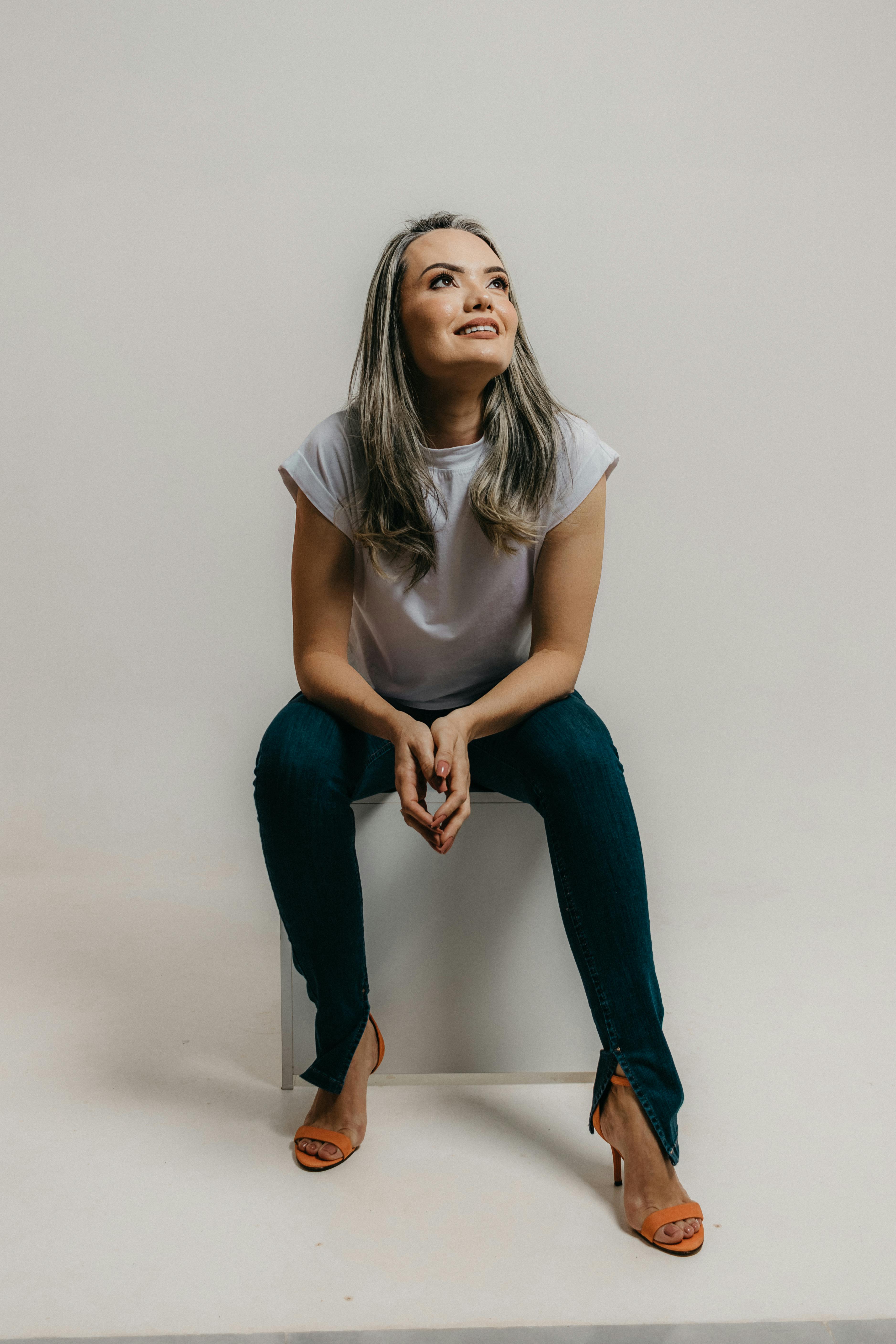 Fashionable woman sitting and posing in a studio, showcasing a modern stylish look with jeans and a white t-shirt.