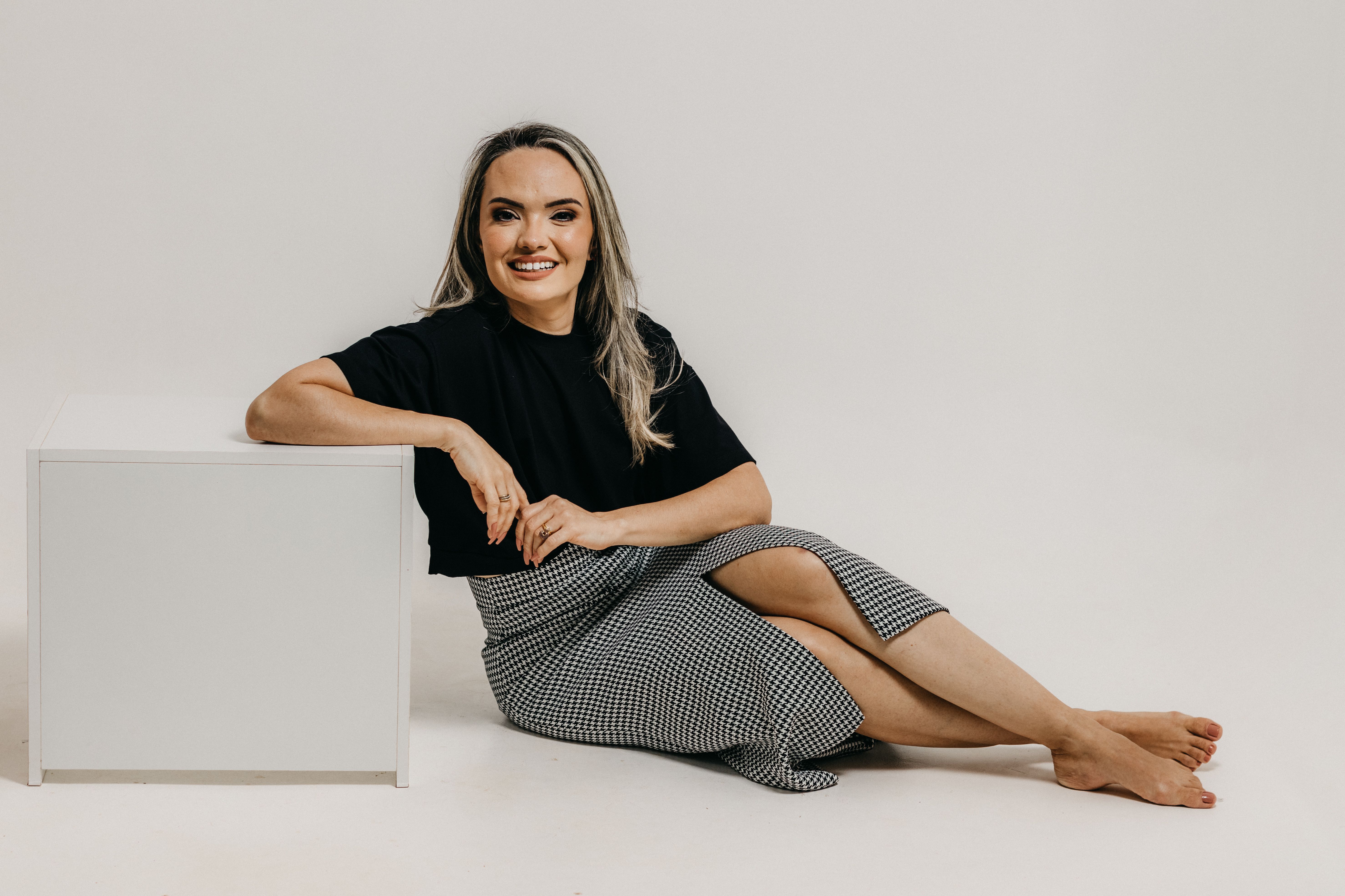 Elegant woman smiling while sitting barefoot on a white background, showcasing modern fashion style.