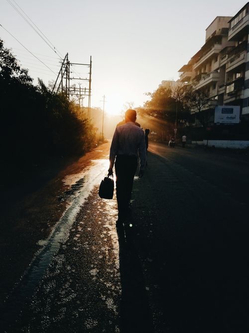 Free A silhouetted man walks along a sunlit street in Pune, India during sunrise, creating a striking urban scene. Stock Photo