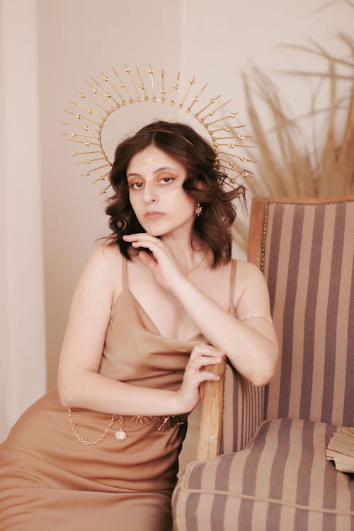 Beautiful Brunette Woman in Light Brown Dress and Golden Sun Rays Headpiece Leaning on Armchair