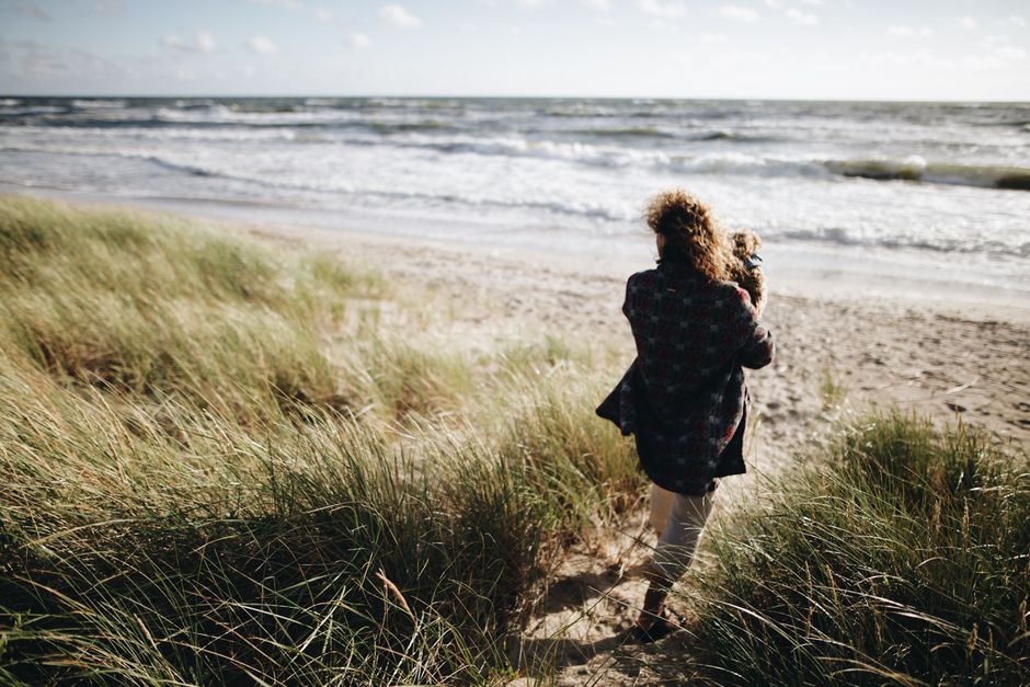 A woman carries a child as she walks towards the serene ocean amidst grassy dunes, capturing a peaceful beach moment.
