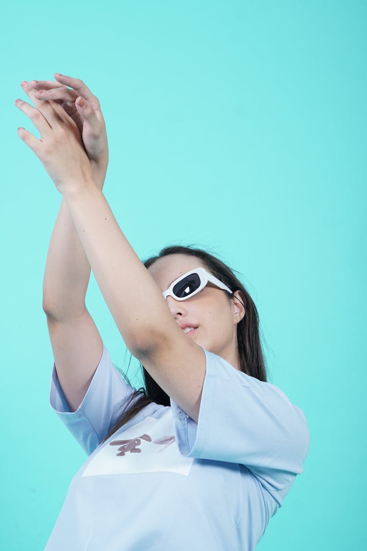 Woman In Sunglasses Standing On Blue Background With Arms Raised In Air