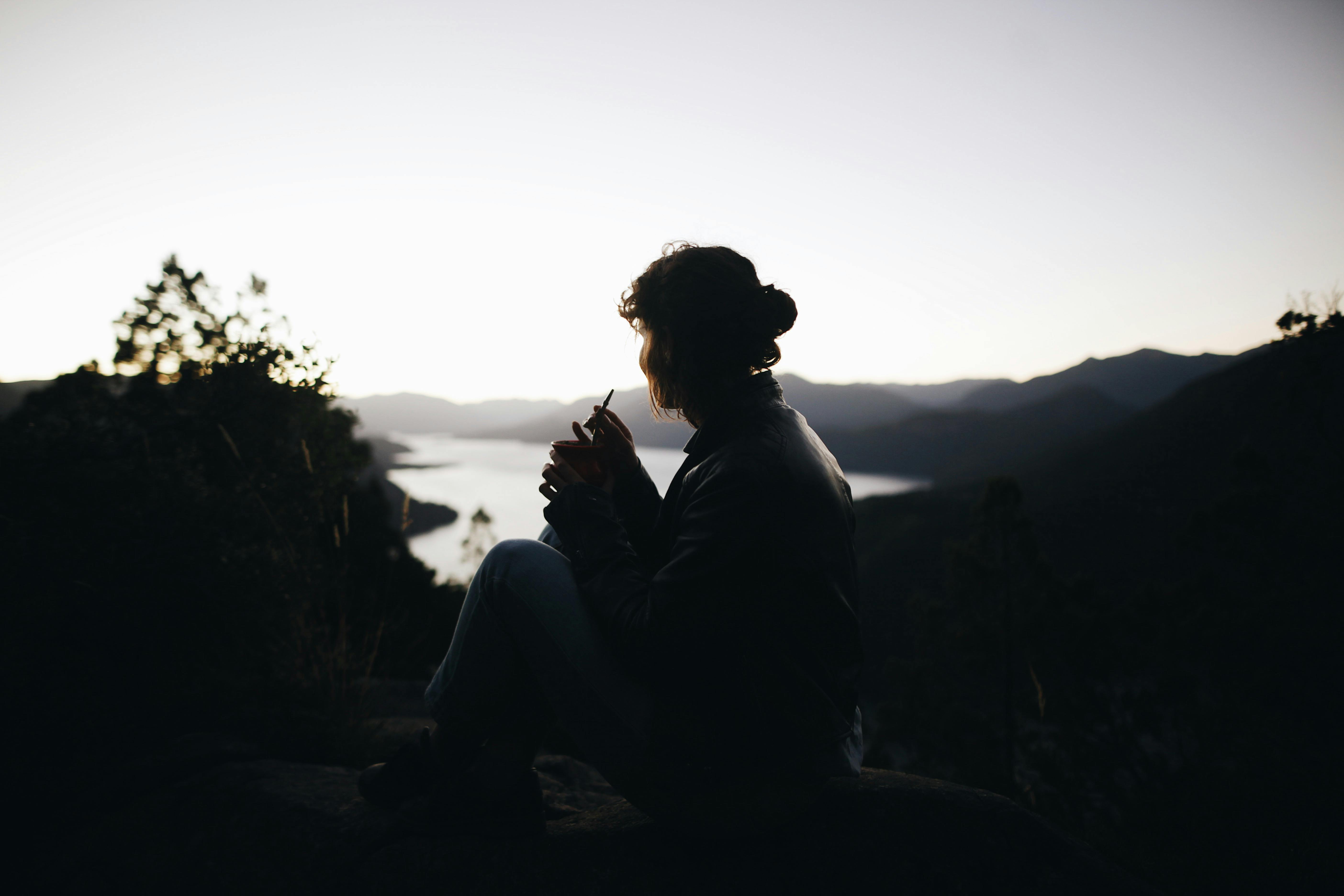 日落时分，一名女子坐在宁静的山间湖畔，身影映照出自然的宁静