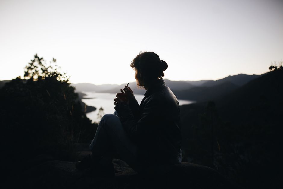 Silhouette of a woman sitting by a tranquil mountain lake during sunset, reflecting serenity and nature.