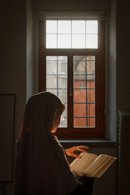 A woman in a hijab reads the Quran near a window in Kayseri, Türkiye.