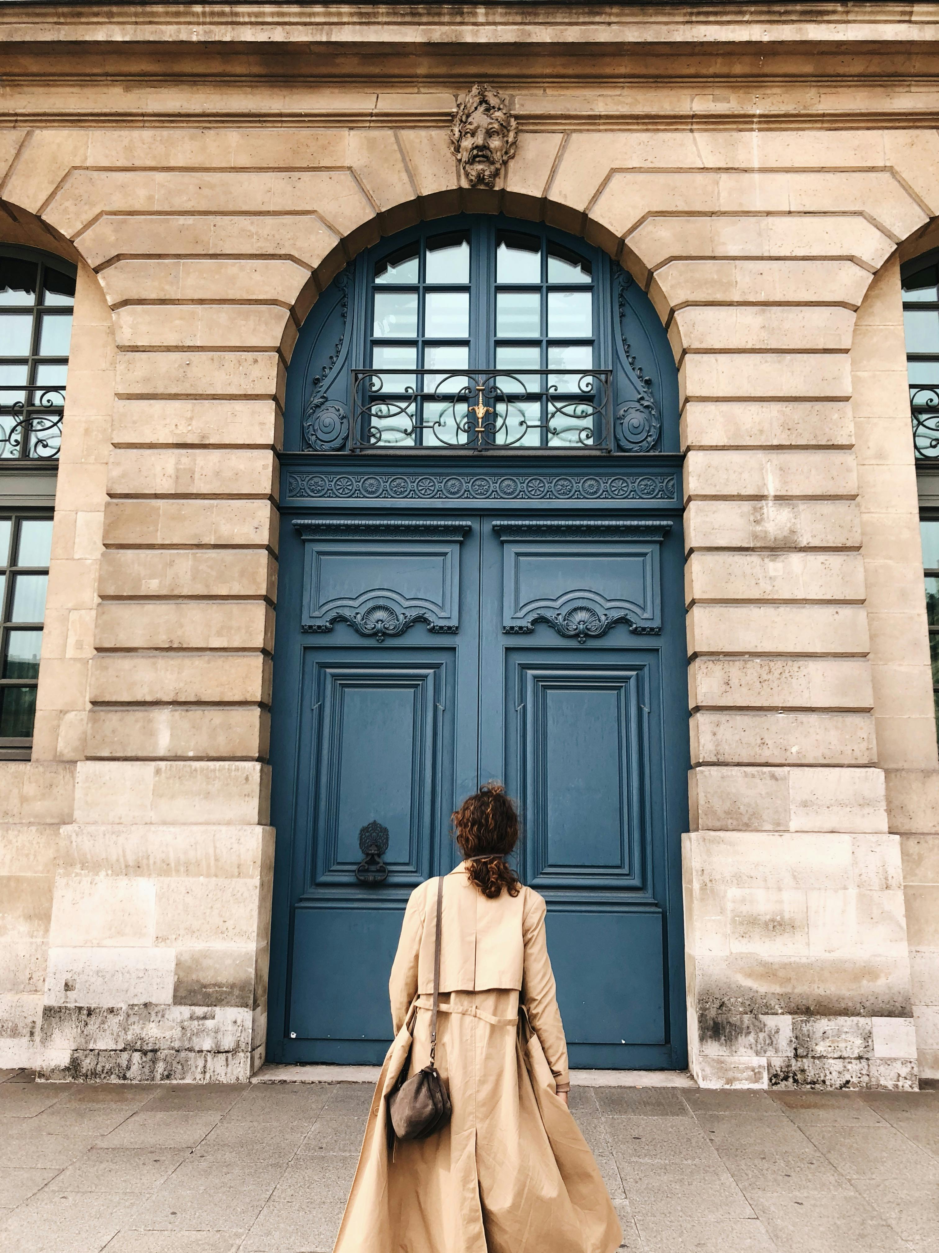 A woman in a trench coat stands before a grand blue door with intricate architecture.