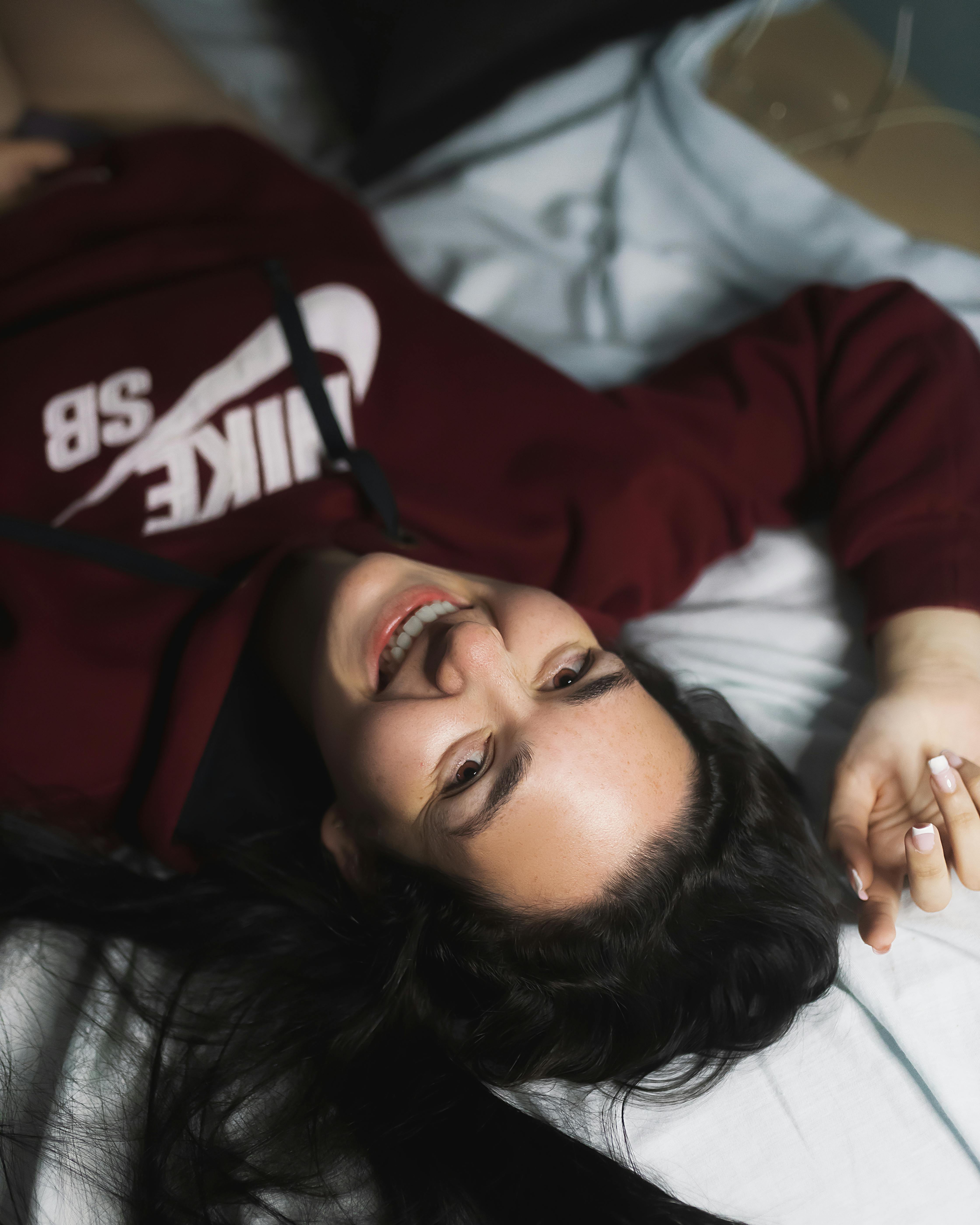 A young woman with black hair lies on a bed, smiling happily, wearing a maroon hoodie.