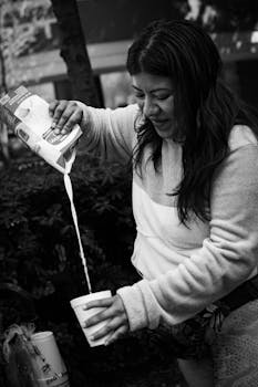 Black and white photo of a woman pouring milk into a disposable coffee cup outdoors.