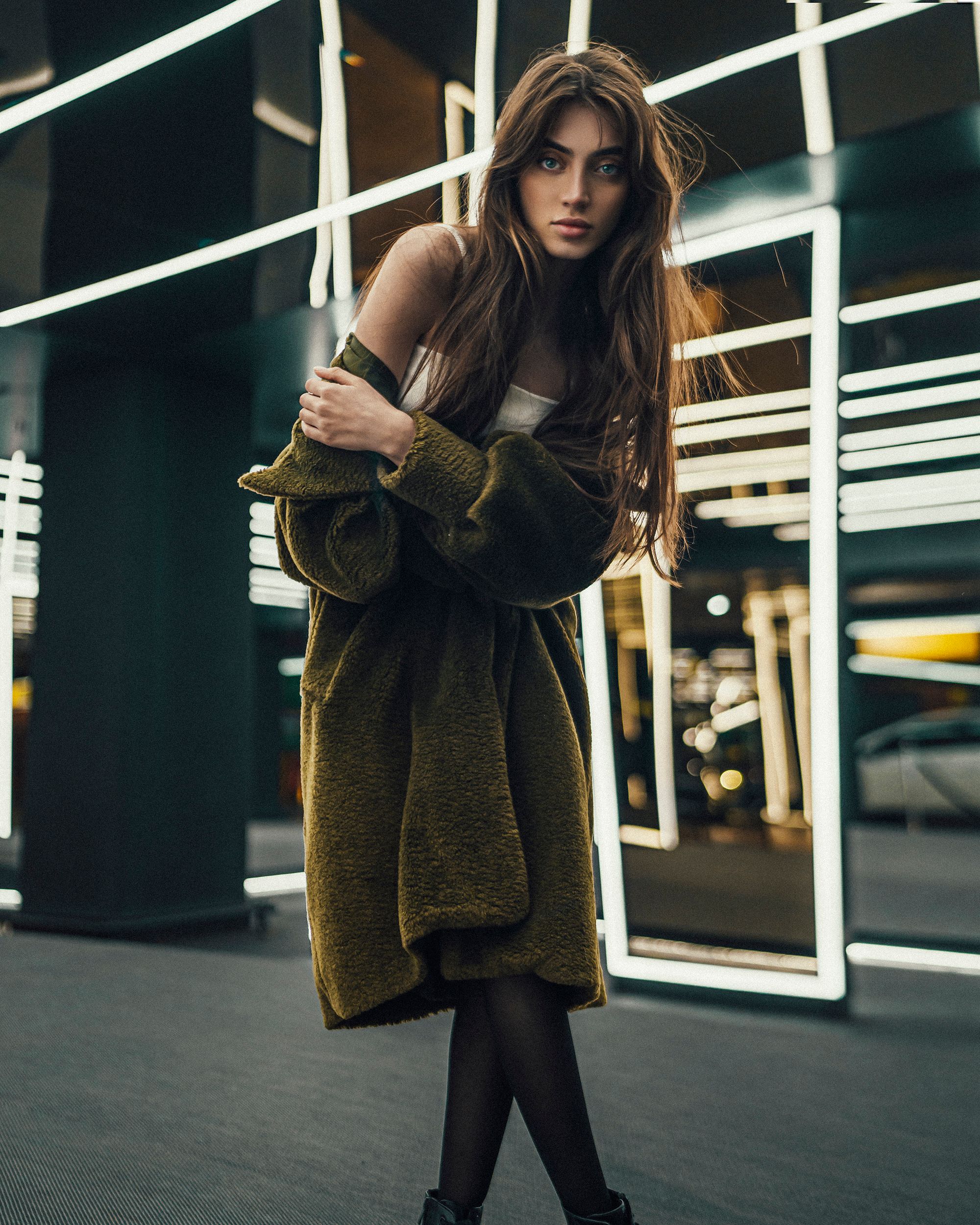 Chic young woman in green coat posing in a modern interior with illuminated mirrors in Istanbul.