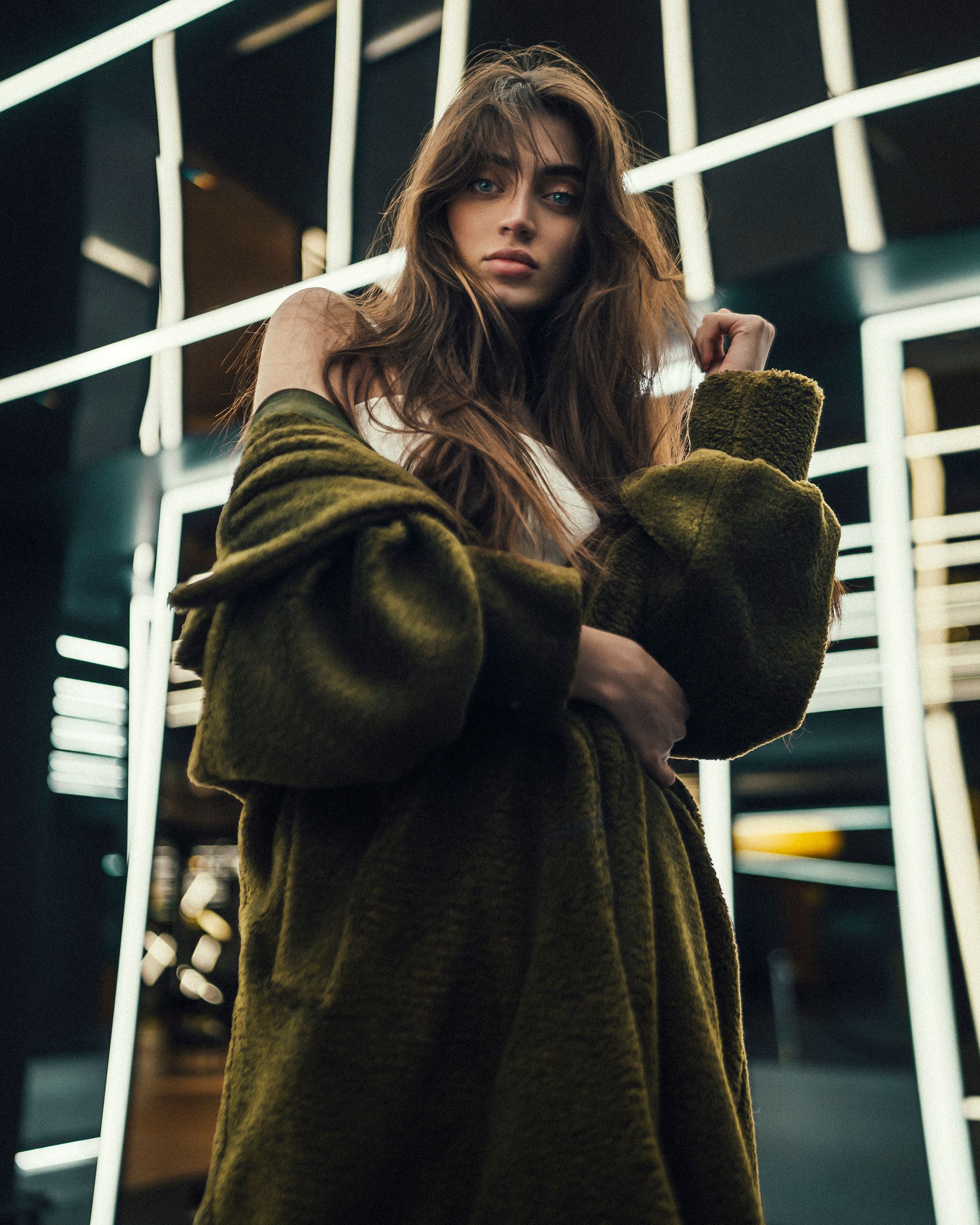 Portrait of a stylish young woman with long brown hair in a trendy green coat, set against a modern backdrop with geometric lights.