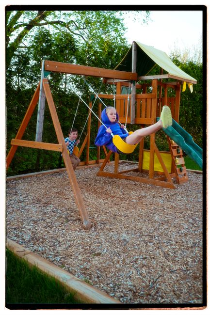 Two children playing on a wooden swing set in a sunny backyard.