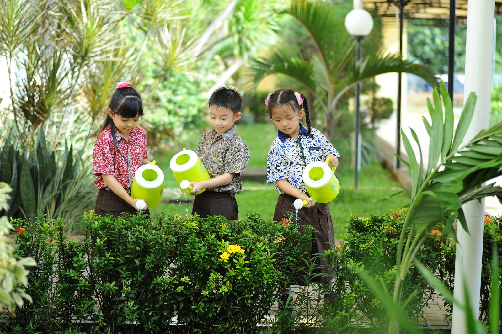 Three young children in school uniforms watering a flower bed with bright green watering cans