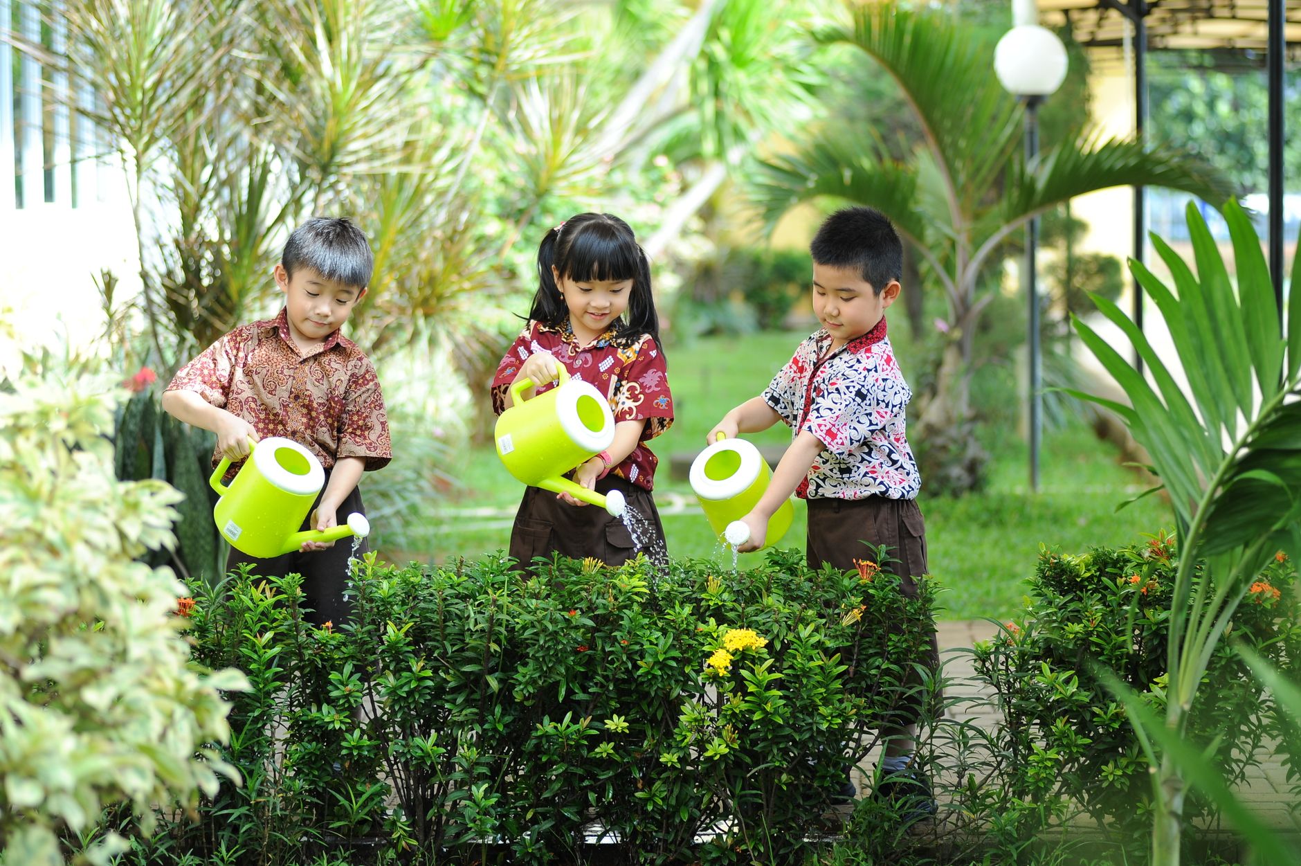 Three children watering plants with bright green watering cans in a vibrant garden setting.