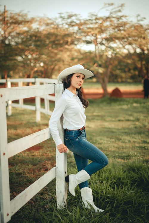 Free Woman wearing a cowboy hat and boots, leaning on a white fence with a rural backdrop. Stock Photo