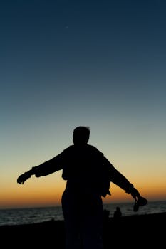 A person in silhouette with arms spread wide at sunset by the sea, evoking freedom and tranquility.