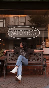 Woman in jeans and boots sitting outside Salon 607 on an ornate bench.