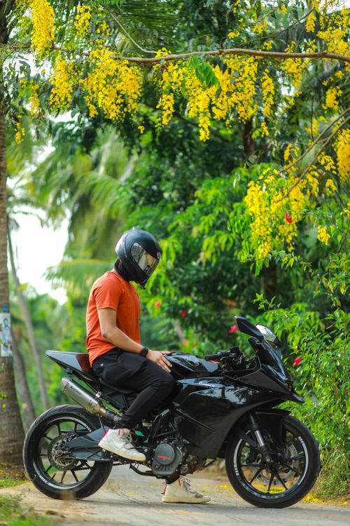 Free A motorcyclist in a helmet sits on a parked bike under a vibrant yellow flowering tree on a scenic country road. Stock Photo