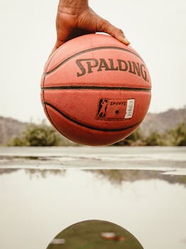 A close-up shot of a hand holding a basketball outdoors near a reflective puddle.