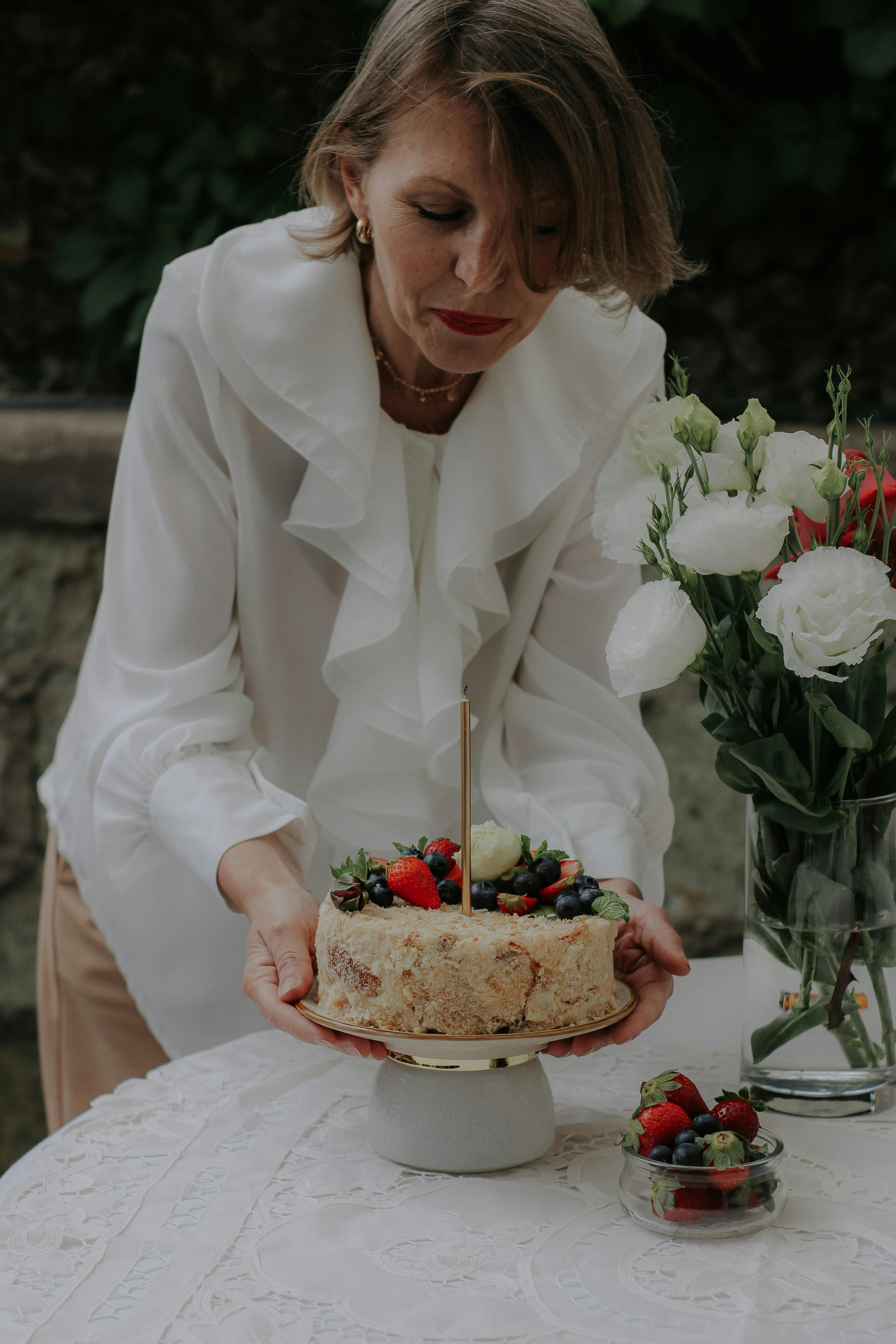 Mature woman in stylish attire placing a berry-topped cake on a table.
