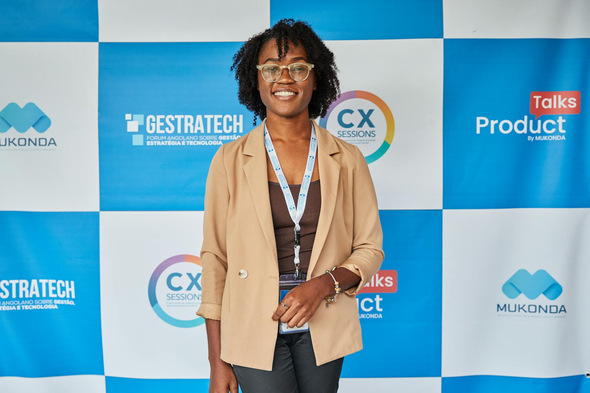 Confident businesswoman in glasses smiling at a tech conference with a promotional backdrop.