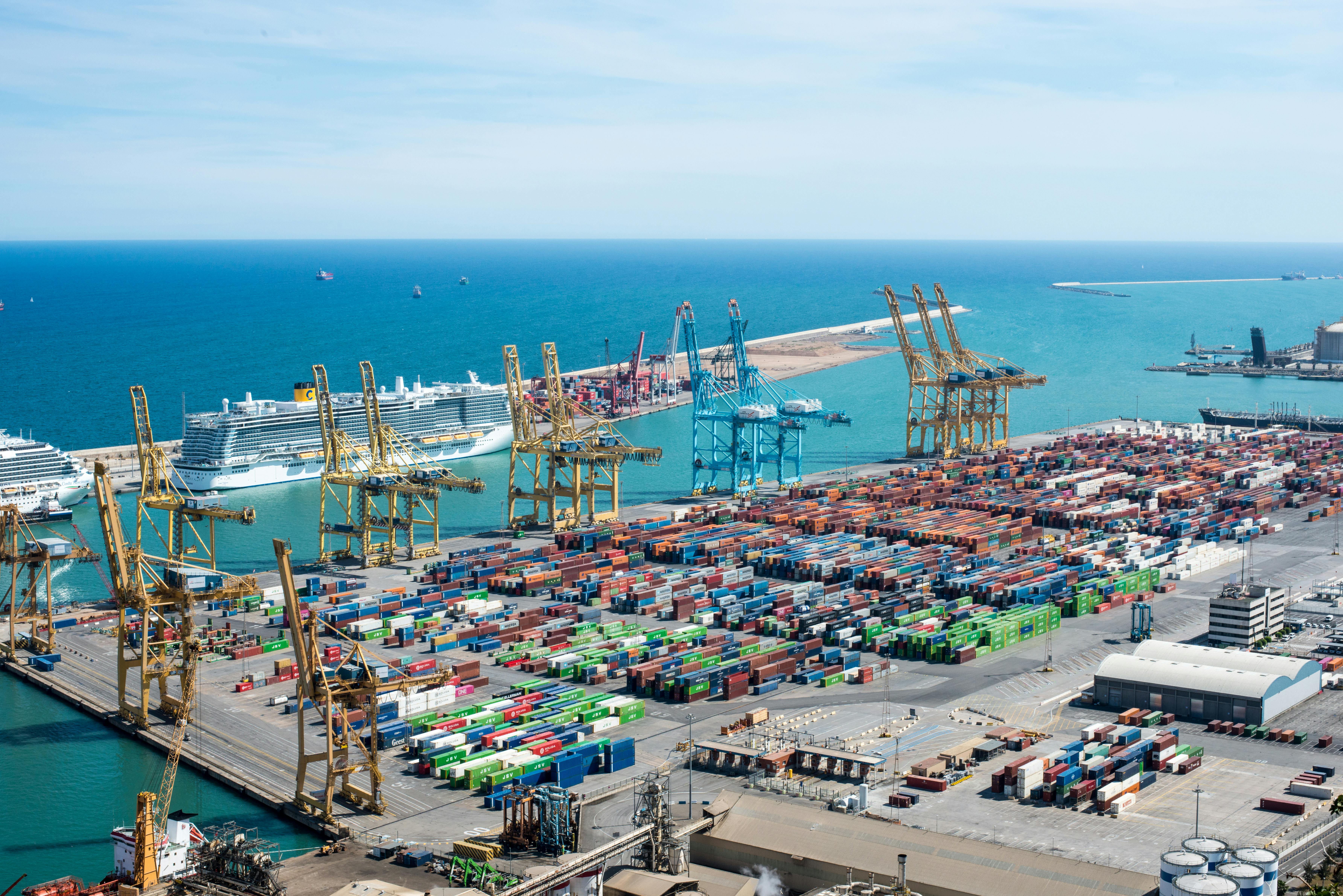 A busy cargo port with stacked shipping containers, large cranes, and cruise ships by the blue waterfront.