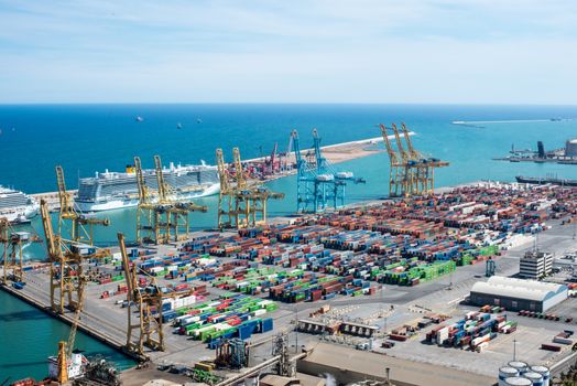 A vibrant aerial shot of Barcelona port featuring cranes, containers, and cruise ships on a sunny day.