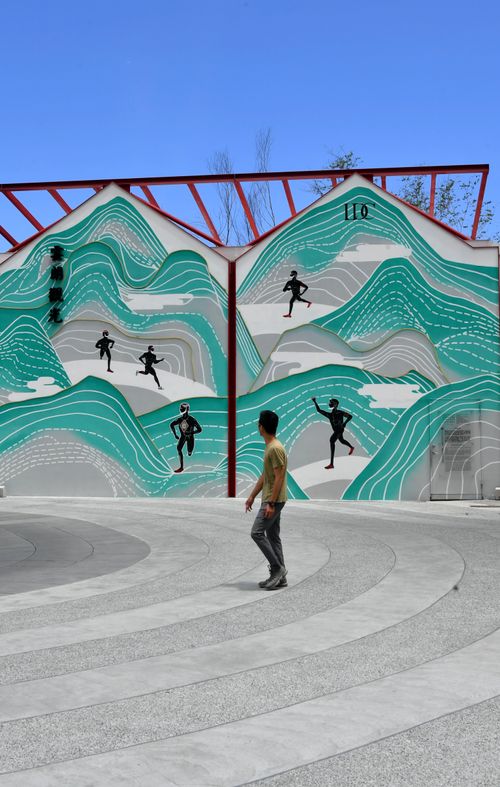 Free A man walking past a vibrant mural depicting athletes on a sunny day in an urban area. Stock Photo