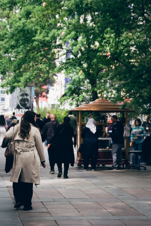 Free Dynamic city street with people walking under lush green trees, vibrant urban life captured. Stock Photo
