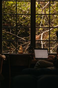 A person working on a laptop by a window with greenery outside. Indoor setting with nature.