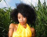 这幅优雅的肖像描绘了一位身着亮黄色服装、留着自然非洲式发型的女性，她正安坐在高高的草丛中