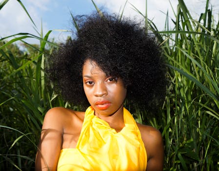 这幅优雅的肖像描绘了一位身着亮黄色服装、留着自然非洲式发型的女性，她正安坐在高高的草丛中