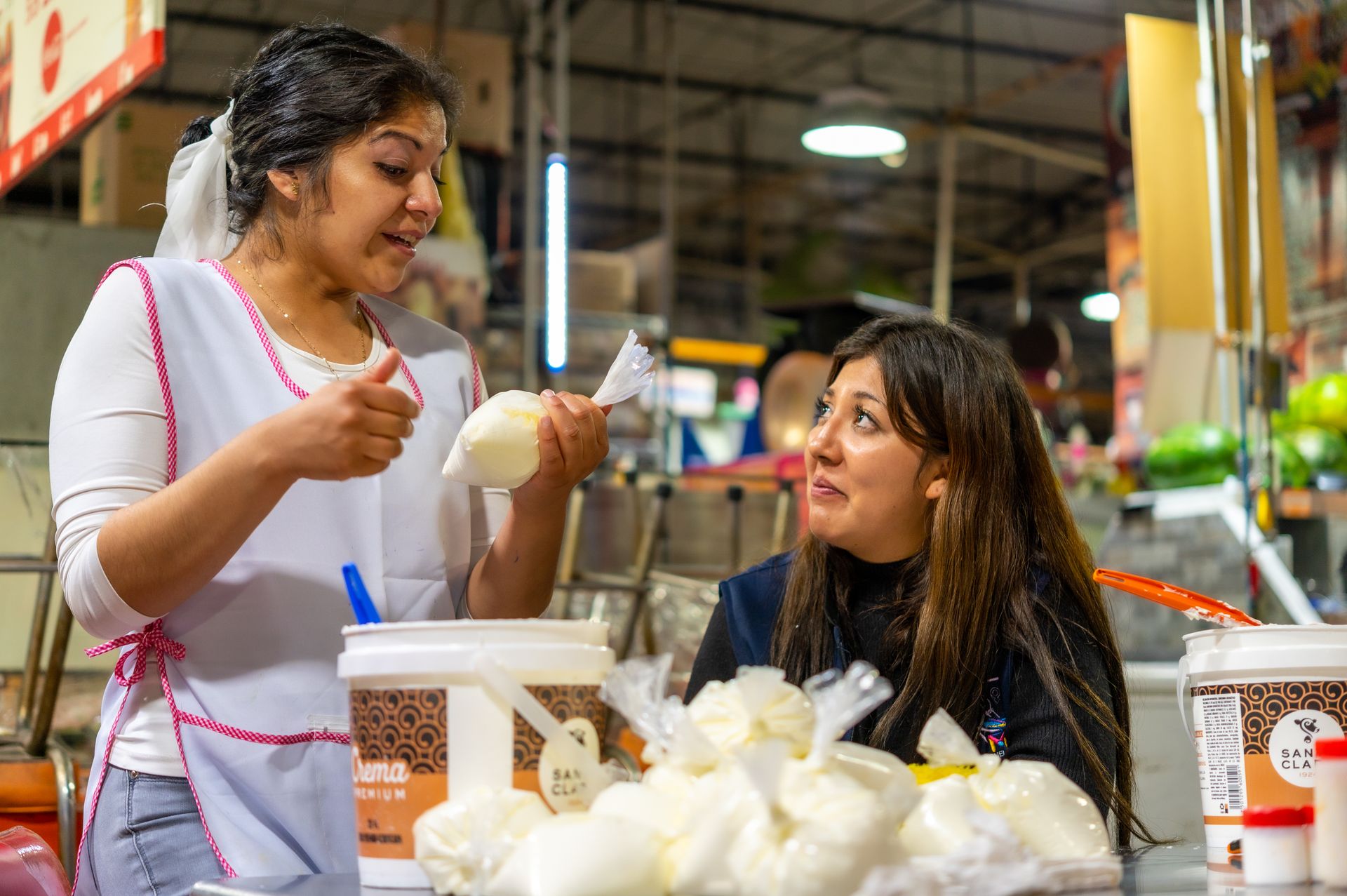 Two women interacting at a vibrant food market, surrounded by dairy products and produce.