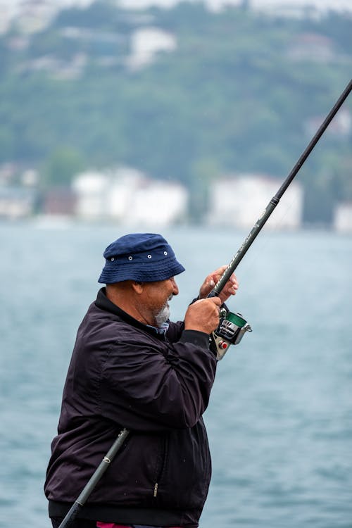 Man in Brown Leather Jacket Wearing Brown Straw Hat Standing on a Dock  while Holding a Fishing Rod · Free Stock Photo