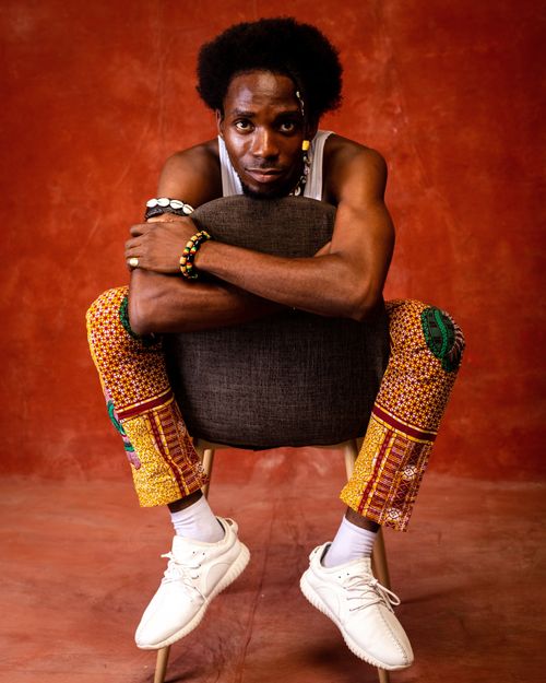 Free Man in vibrant attire posing with chair against orange backdrop, showcasing unique fashion style. Stock Photo