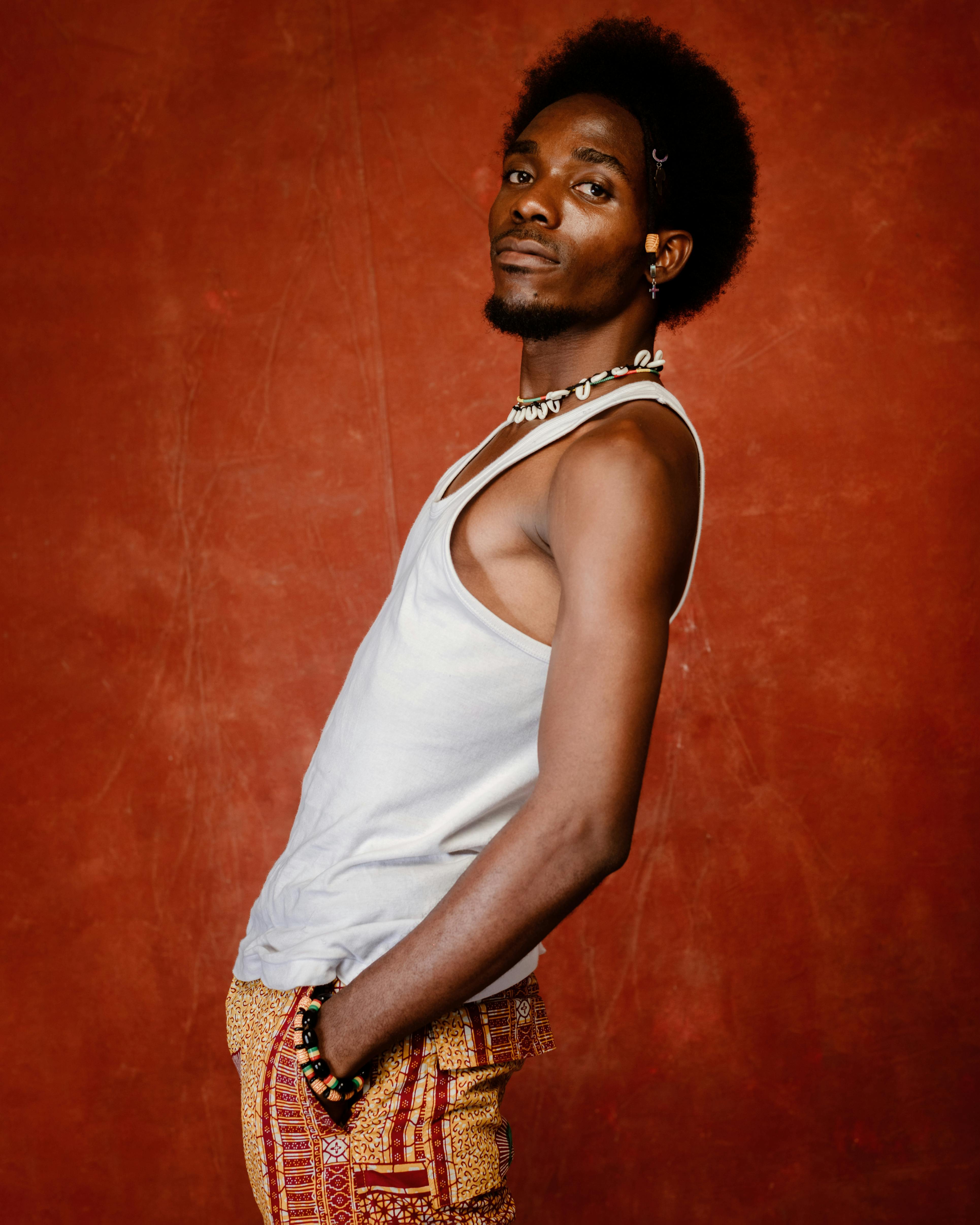 Artistic portrait of a stylish young man in a tank top and patterned pants posing in a studio setting with a warm background.